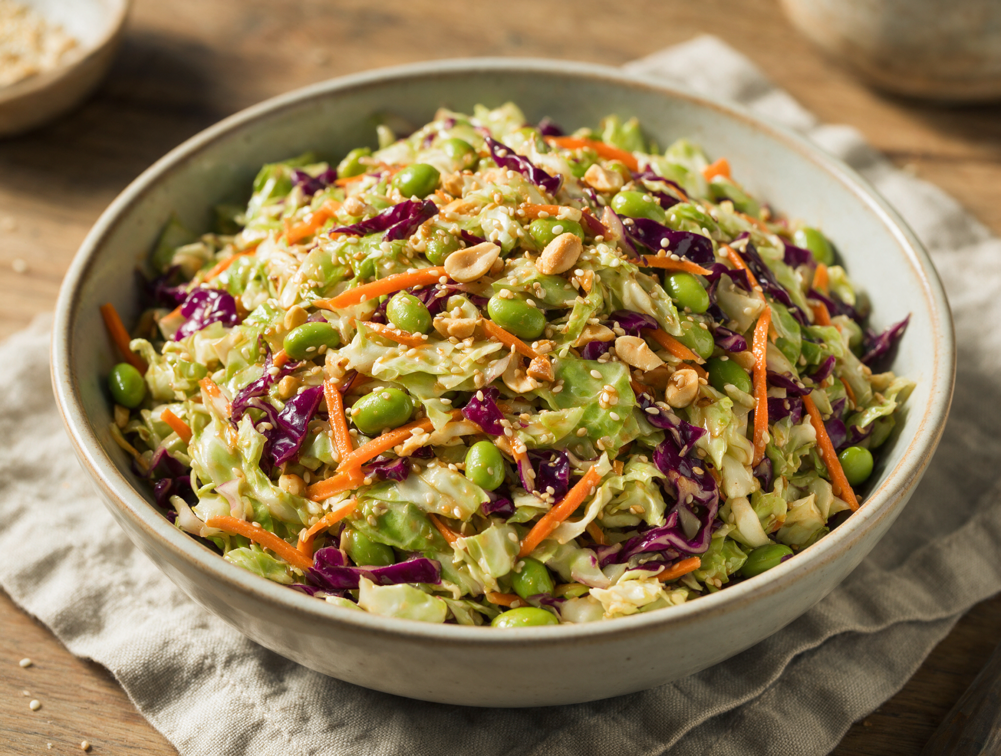 Wide bowl of shredded cabbage slaw topped with edamame, peanuts and sesame seeds, glossy dressing, shot on a linen napkin.