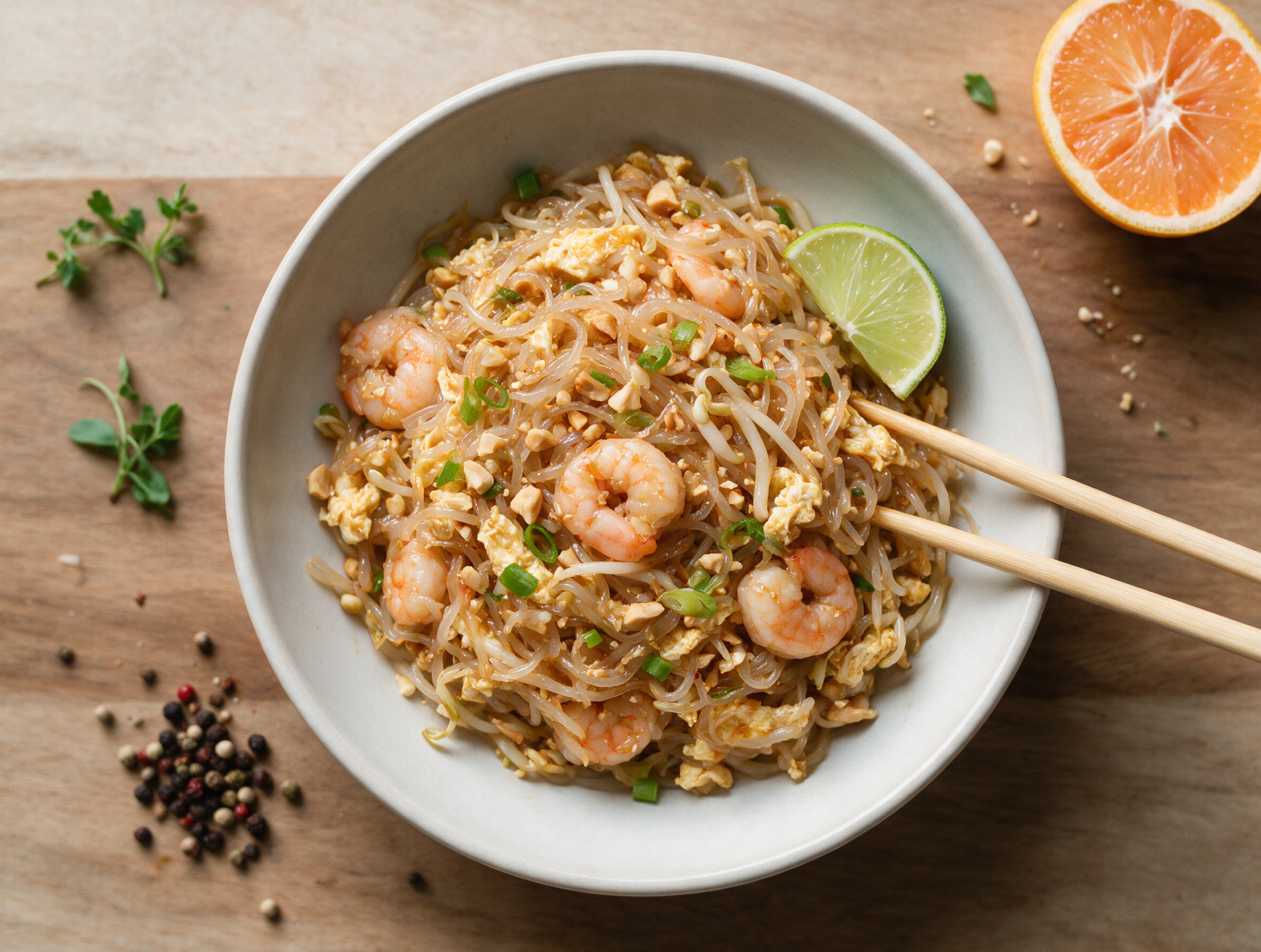 Overhead bowl of shirataki noodle pad thai with pink shrimp, egg ribbons, crushed peanuts and a lime wedge on a wooden table.