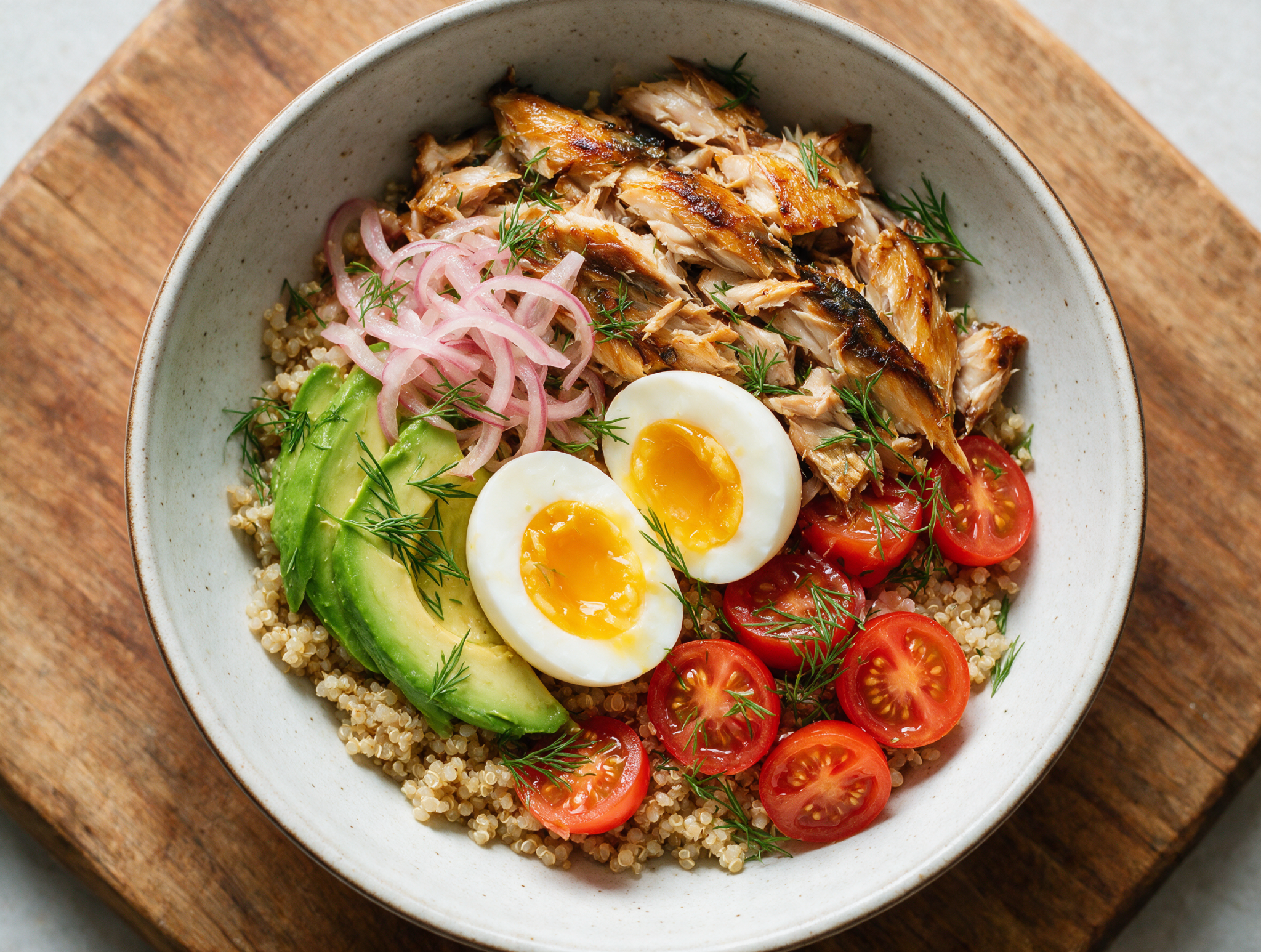 Overhead shot of a smoked mackerel egg bowl with jammy egg halves, avocado slices, and pickled red onion on a wooden board.