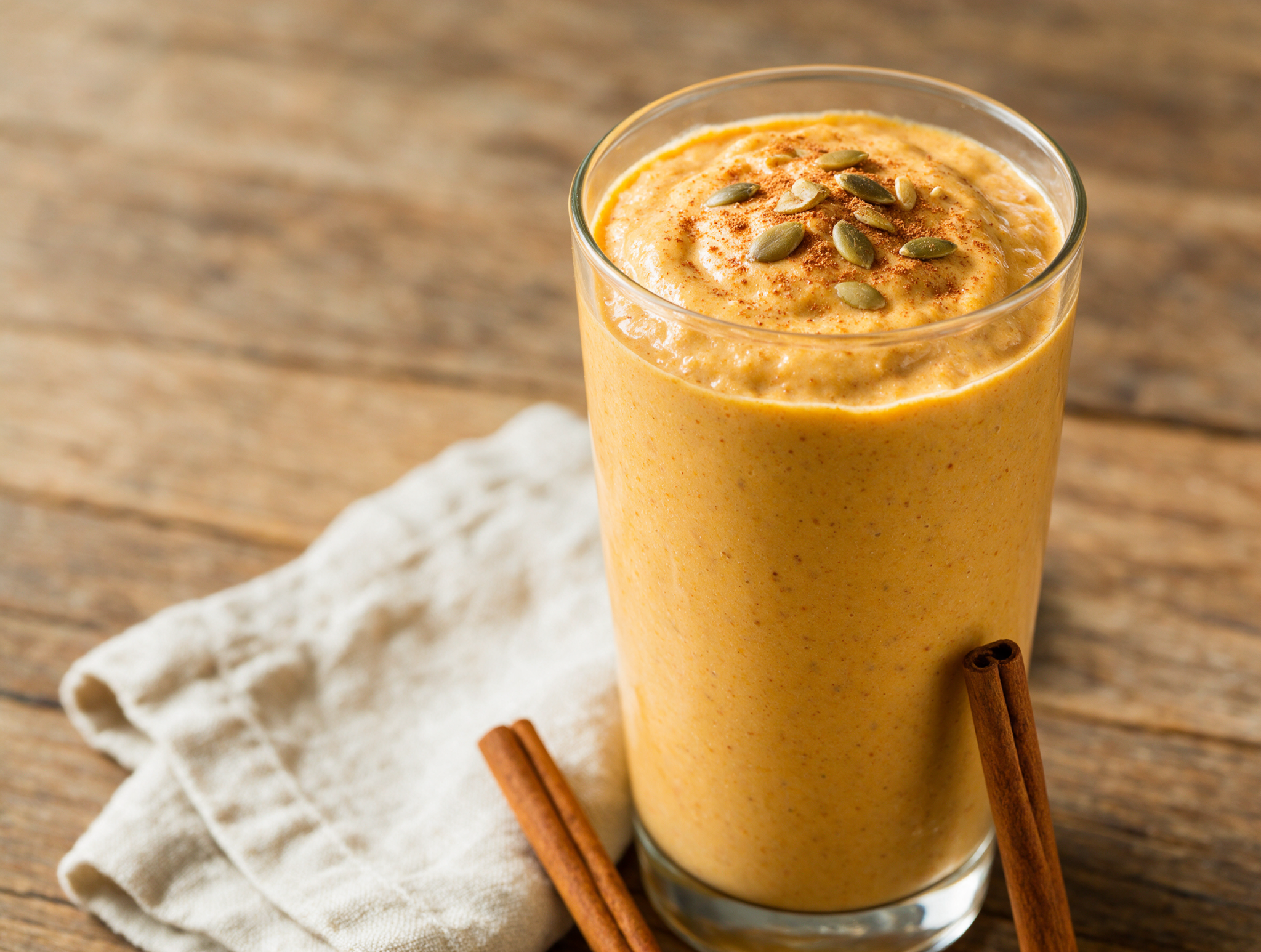 Overhead view of a pumpkin spice protein smoothie in a tall glass with cinnamon dust and pumpkin seeds on a wood board.