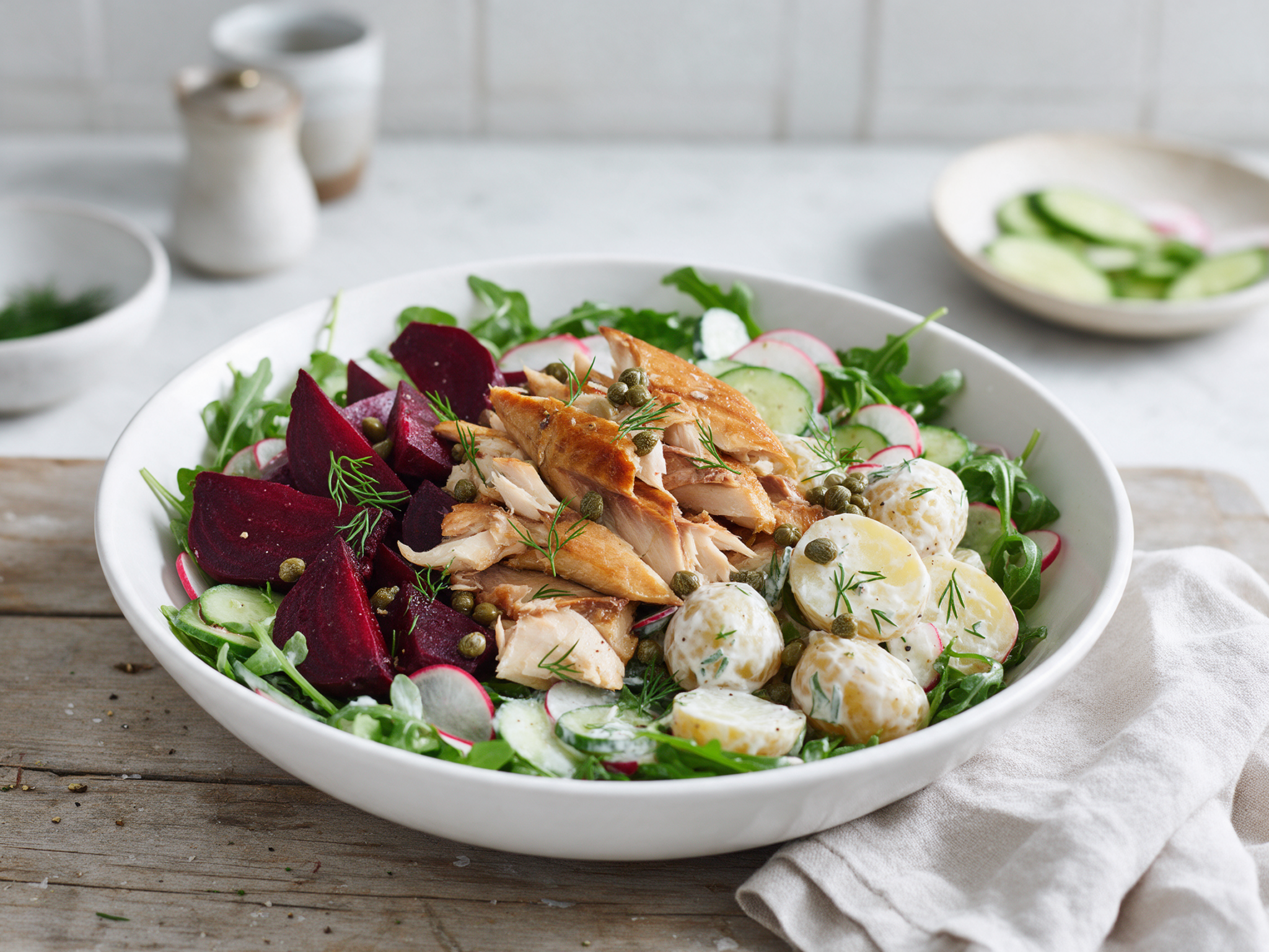 Overhead white bowl of flaked smoked mackerel, ruby beet wedges, potatoes and arugula on a wooden table